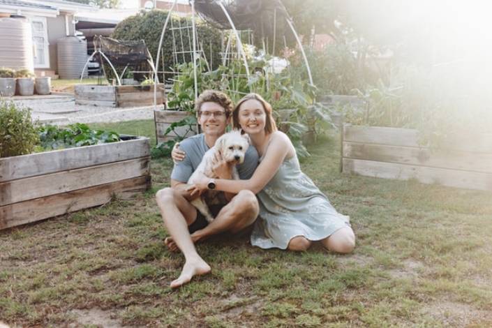 two house sitters cuddling a small white dog in front of a vegetable garden