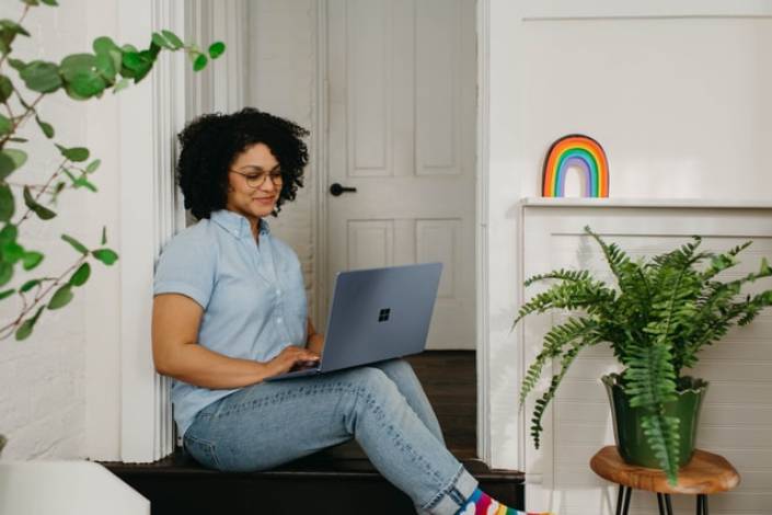house sitter wearing jeans and a blue button up shirt sitting on a step smiling while looking at a computer with a plants and a rainbow painting in the background